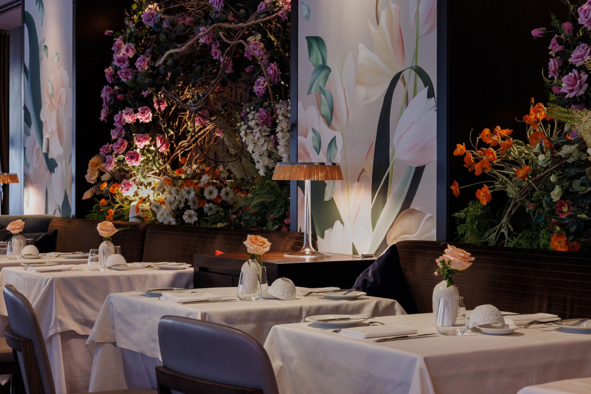 Dimly lit dining room with white-linen tables, rose bud vases, and lush floral murals