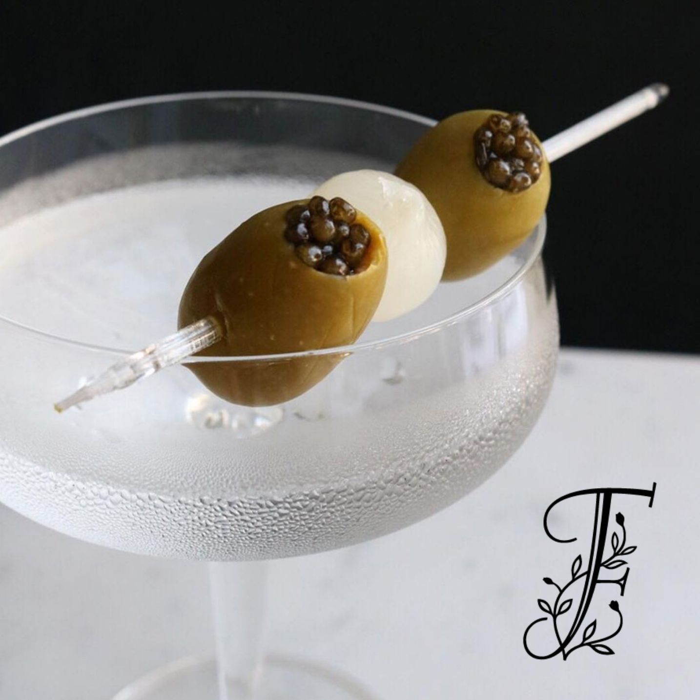 Frosted coupe martini garnished with caviar-topped olives and a cocktail onion on a skewer