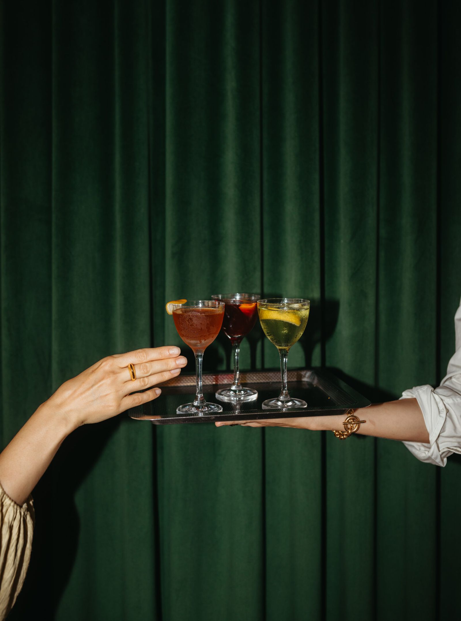 Server's outstretched arm presenting a tray of three citrus-garnished cocktails against green velvet
