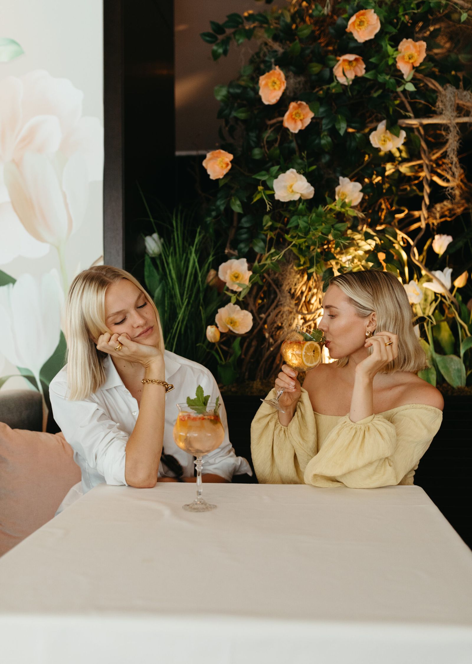 two young woman sip on gin and tonics in goblets in front of a display of flowers