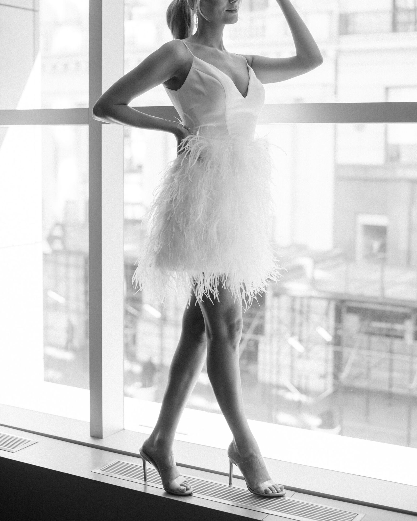 Bride in a short feathered dress posing by floor-to-ceiling windows overlooking the city