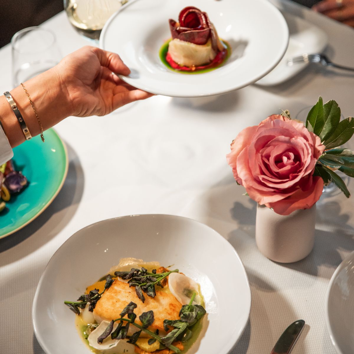 Hand placing a plated seafood dish on a white-linen table beside a pink rose in a bud vase