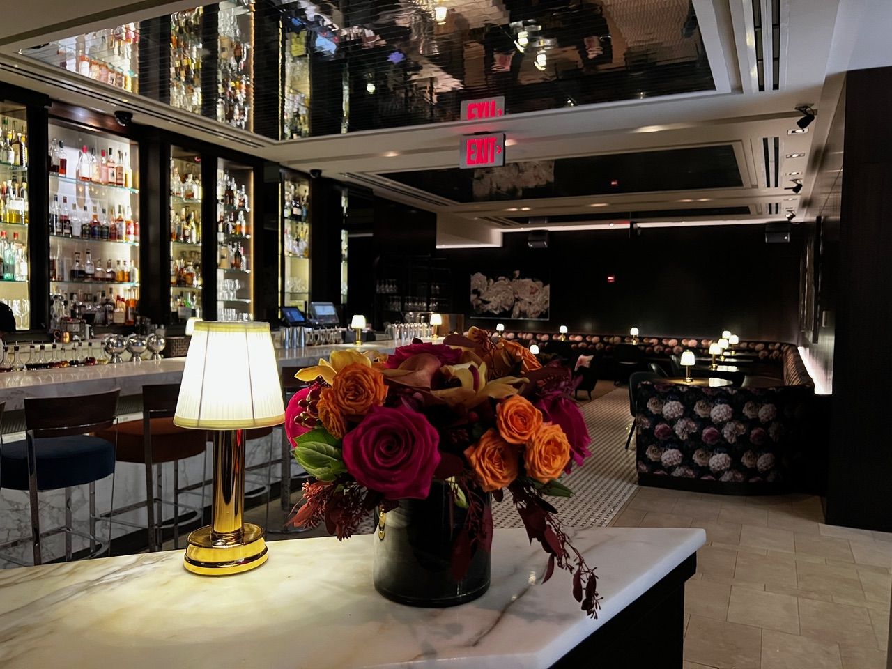 Marble host stand with a brass lamp and rose bouquet overlooking a dimly lit backlit bar