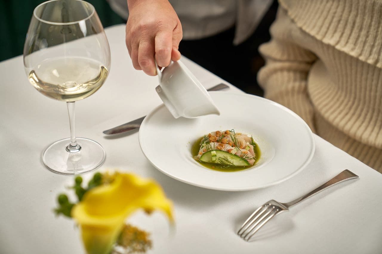 Server pouring green broth over a crudo and cucumber dish beside a glass of white wine