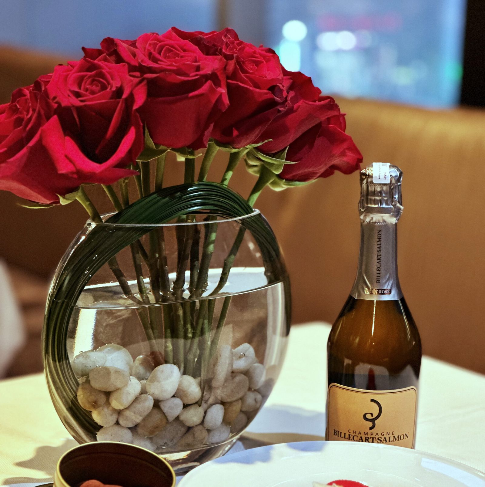 Red roses in a glass bowl vase with white stones beside a split of rosé Champagne