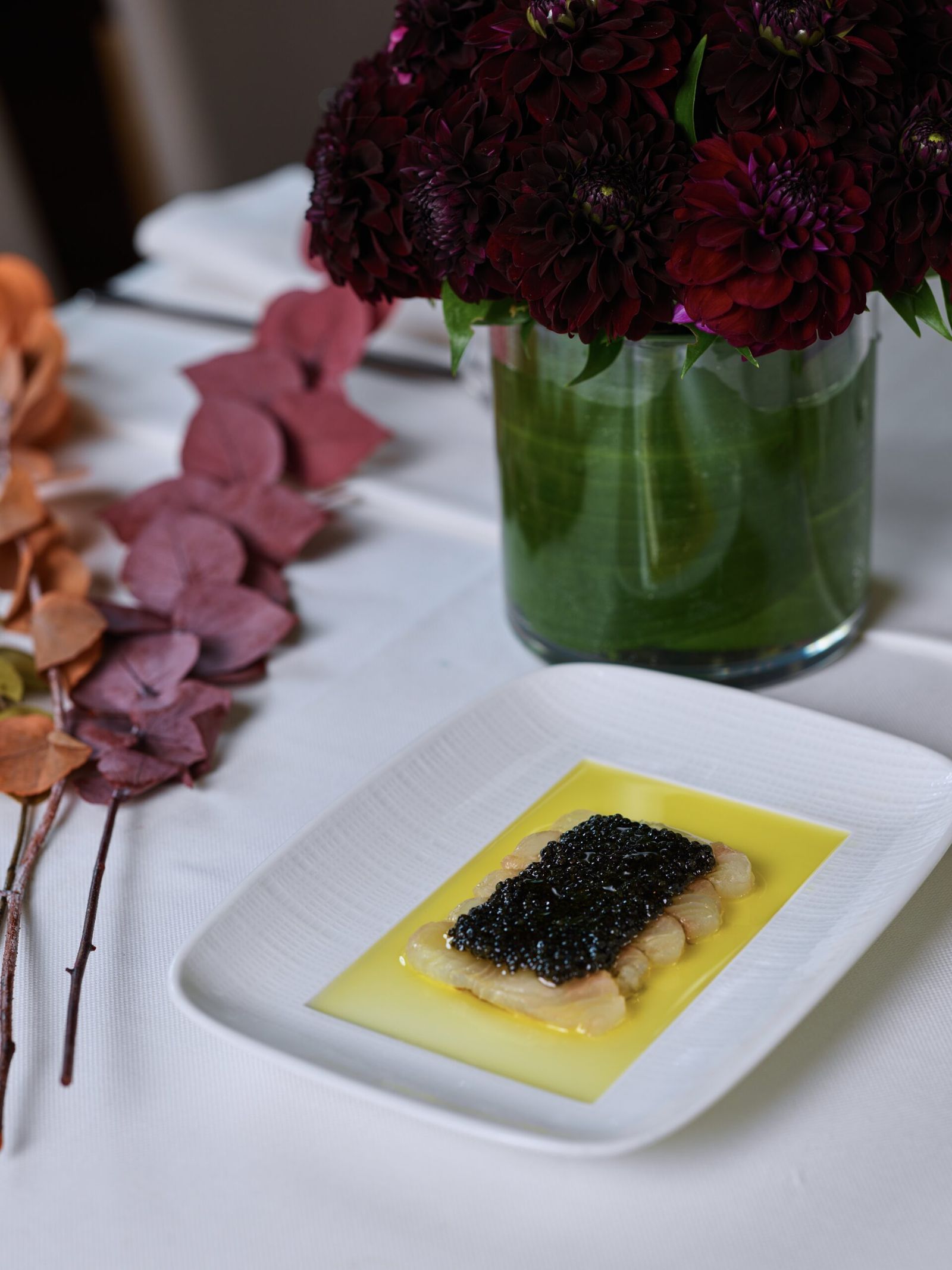 Caviar over thinly sliced fish in golden olive oil on a white plate beside dark dahlias