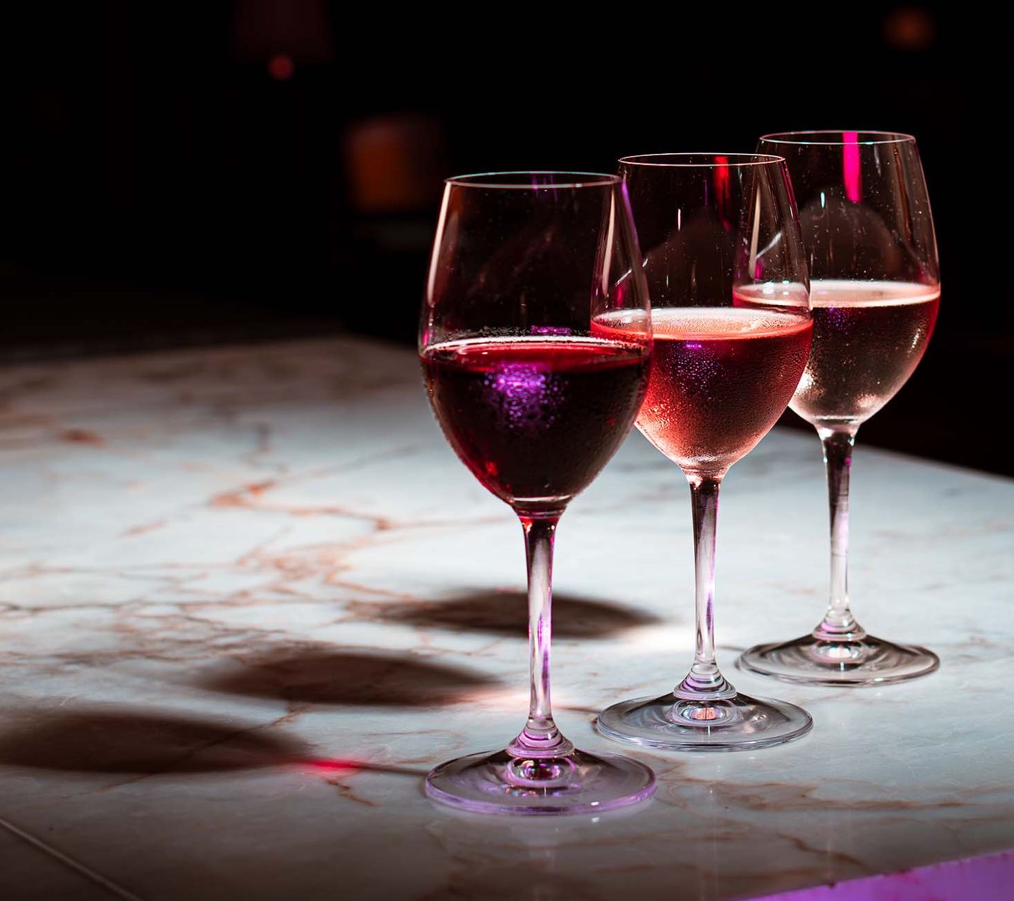 Three glasses of red, rosé and white wine on a marble bar top under moody pink lighting