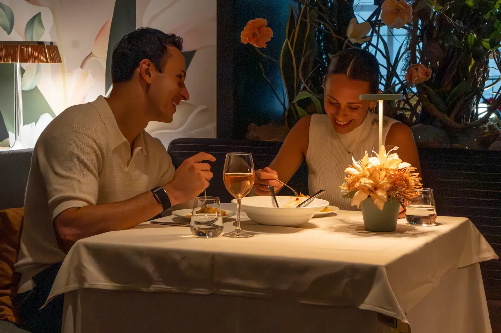 Smiling couple sharing a candlelit dinner and wine at a white-linen table with floral décor