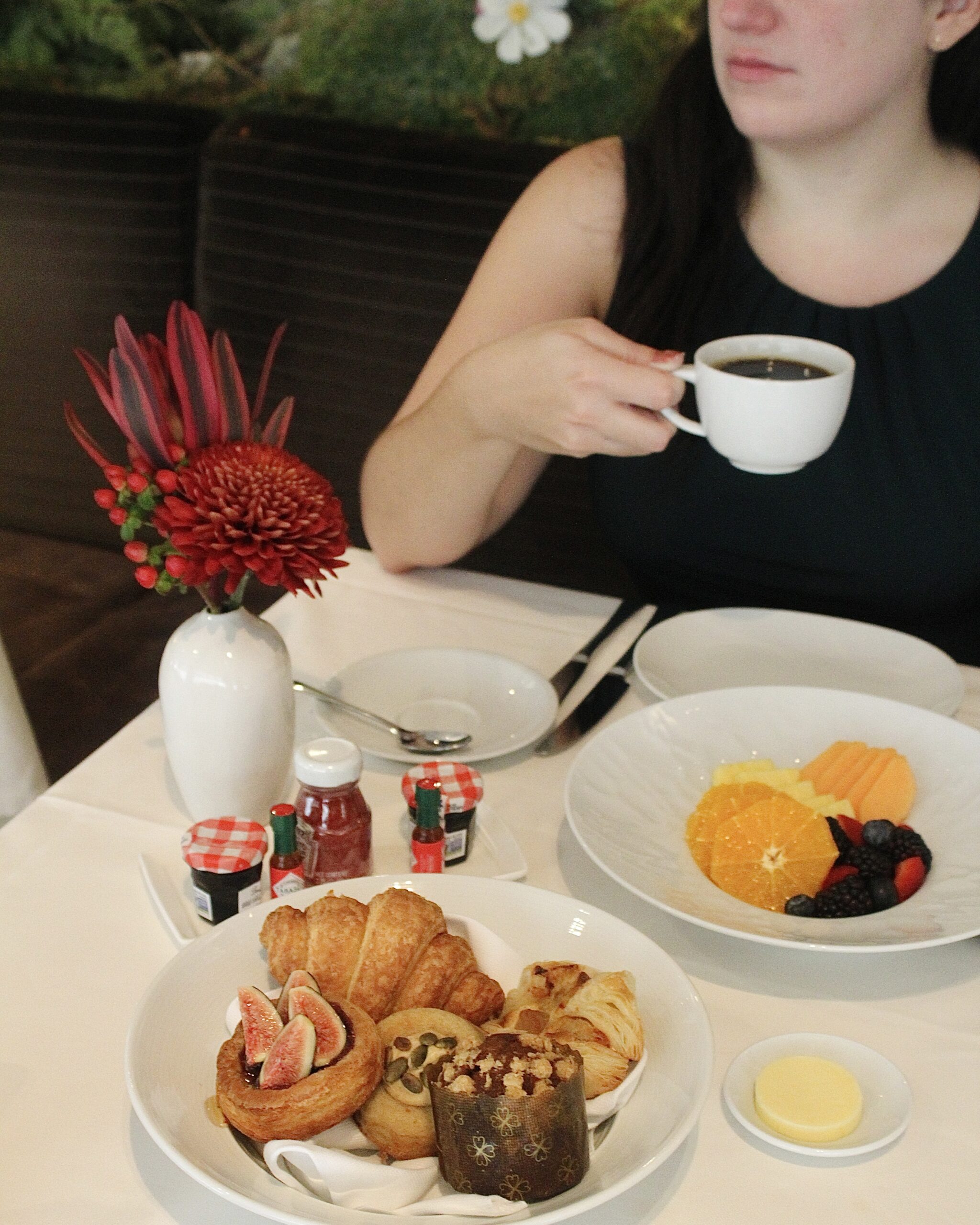 a woman drinks a cup of coffee at a table full of breakfast items: a bowl of fruit and a basket of pastries.