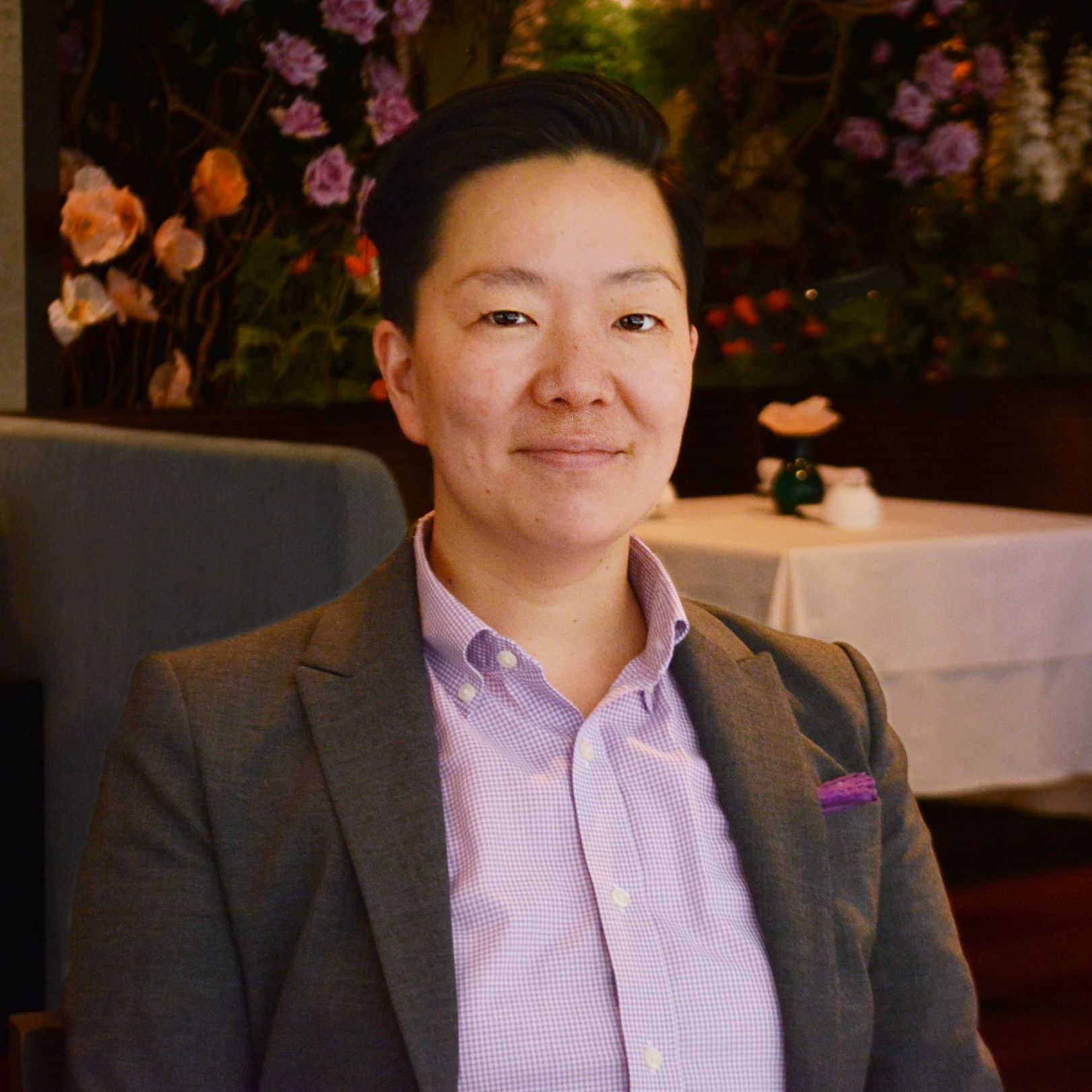 susan lee, a woman with short black hair, wearing a lavendar button down under a grey blazer. in the background we see the ai fiori dining room - tables with white table cloths and flowers.