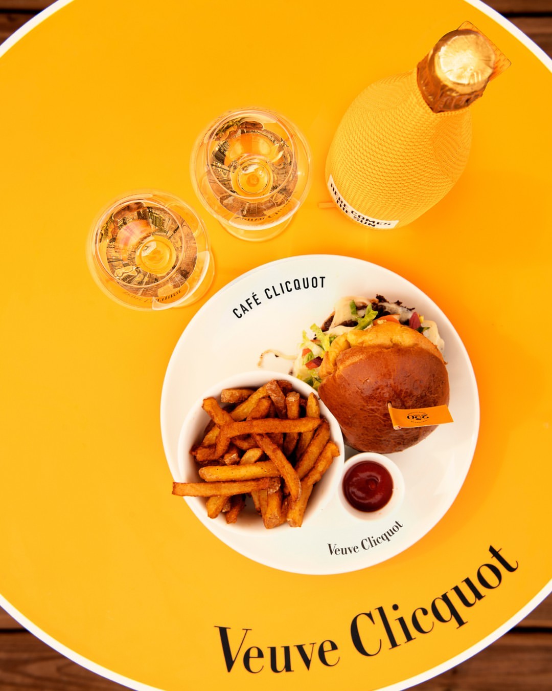 a top down photo of a burger and fries with two glasses of veuve cliquot champagne, and a bottle of champagne. all sitting on a bright yellow-y orange table - veuve branded.