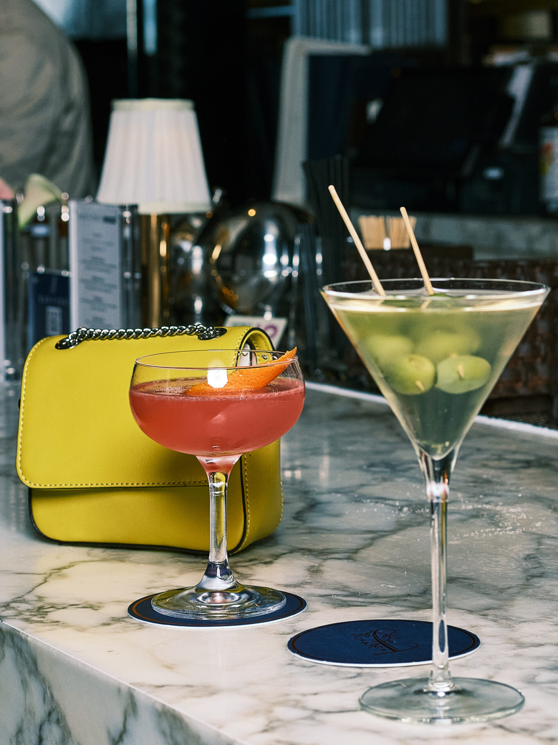 a yellow purse sits on a marble bar, behind a coupe glass with a pink cocktail and a dirty martini with olives