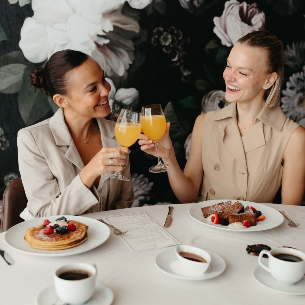 two women toast mimosas over a breakfast of pancakes and french toast