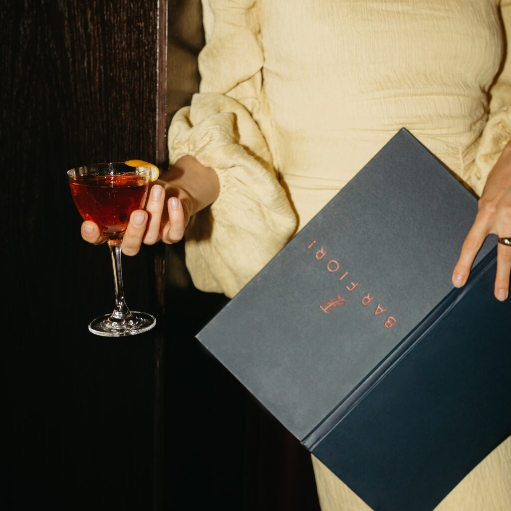 an elegantly dressed woman holds a bar fiori menu in one hand and a $14 martini in the other