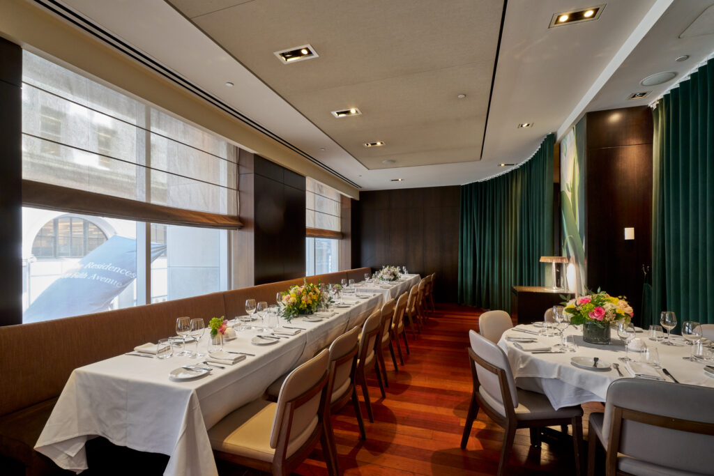 ai fiori's la spezia semi-private dining room and event space. a room with large windows to the left, and a mix of wooden walls and emerald curtains to the right. along the windows is a brown banquet and a long table with a white tablecloth, fully set. to the right is a large round table, also fully set.