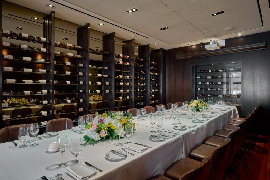ai fiori's private dining room, the monterosso wine room. a long table with a white table cloth, fully set and with vibrant flowers, is surrounded by wine racks.