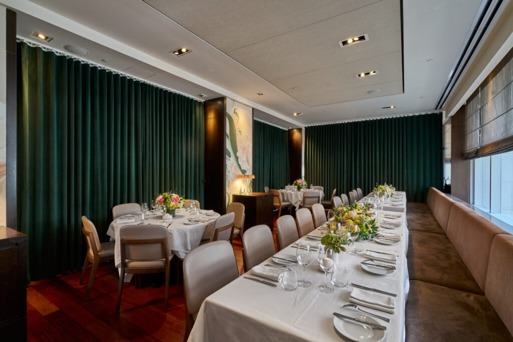 ai fiori's genoa semi-private dining room and event space. a room with large windows to the right, and a mix of floral wallpapered walls and emerald curtains to the left. along the windows is a brown banquet and a long table with a white tablecloth, fully set. to the right is two large round tables, also fully set.