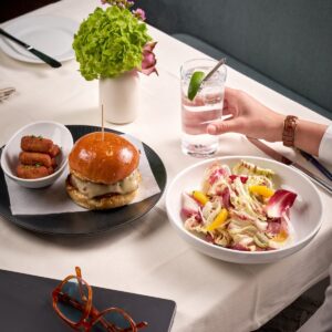 Elegant business lunch setup at Ai Fiori featuring a gourmet burger with croquettes, a fresh citrus radicchio salad, and a sparkling cocktail, on a white tablecloth with a laptop and glasses nearby.