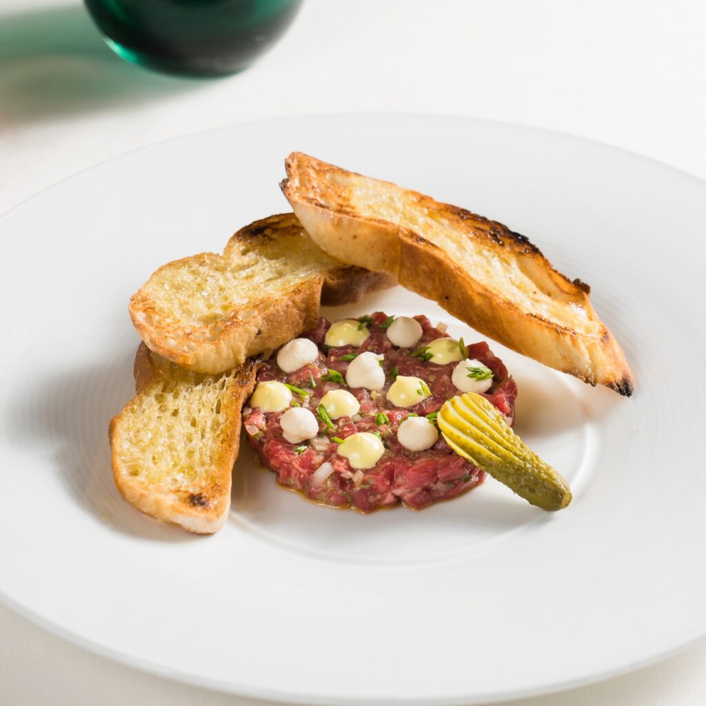 beef tartare with a cornichon and crostini on a white plate