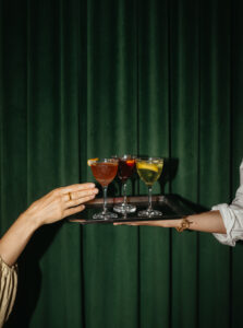 one hand holds a tray with various martinis in nick and nora glasses while another hand reaches to grab one