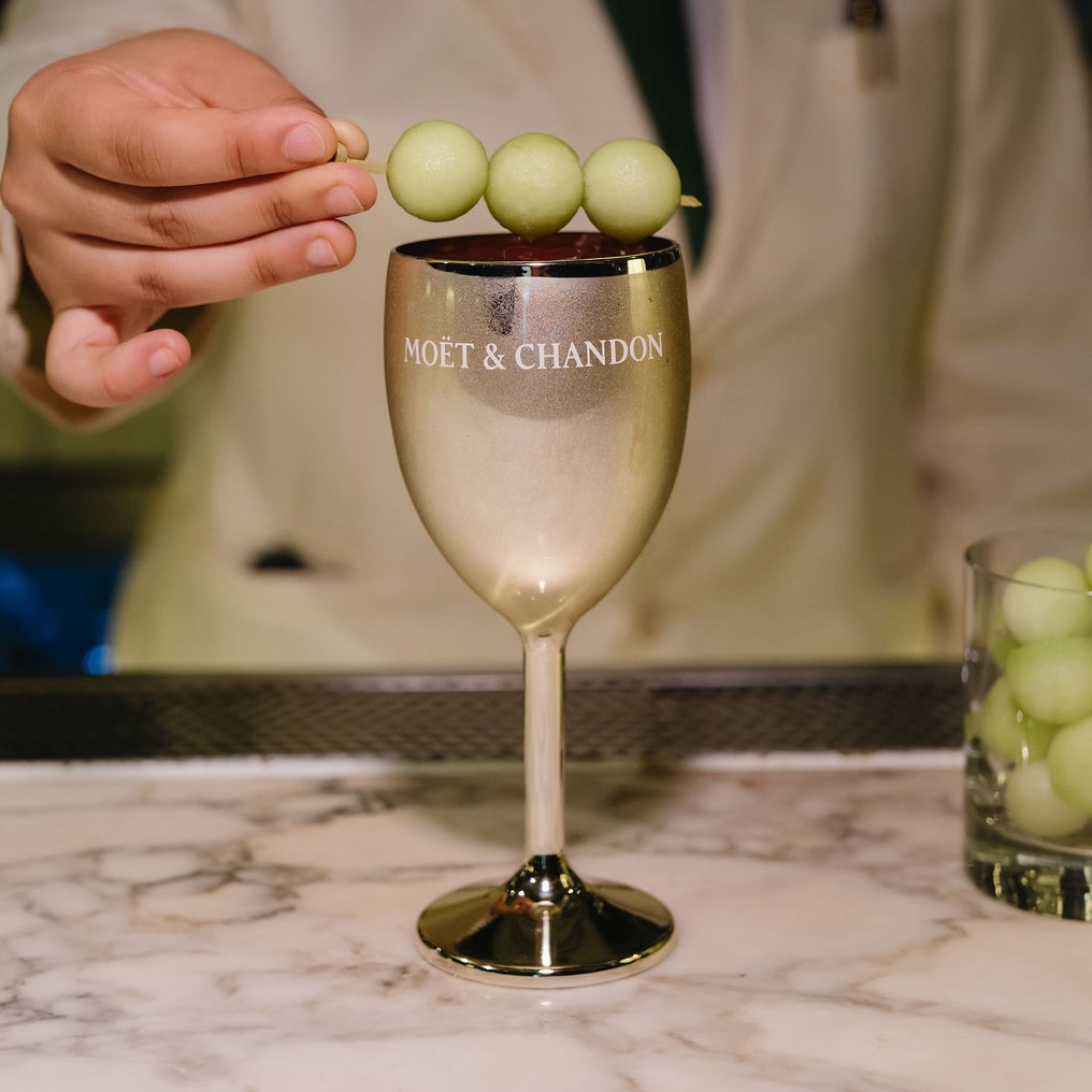 a bartender adds a skewer with 3 balls of honeydew melon to the top of a golden goblet with the moet and chandon logo.