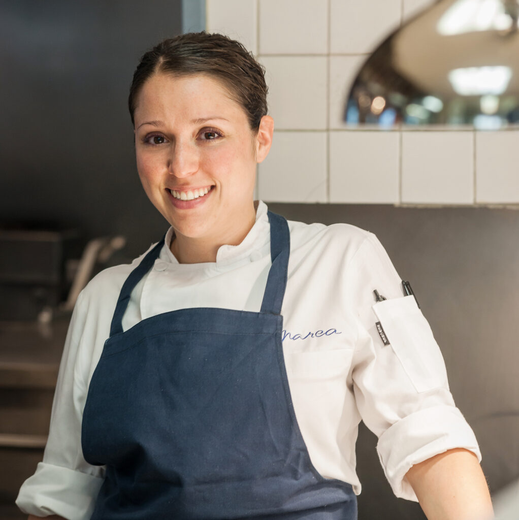 Executive Chef Lauren DeSteno of Ai Fiori, standing in the kitchen with a warm, confident smile, wearing her chef’s jacket and surrounded by the refined atmosphere of the restaurant.